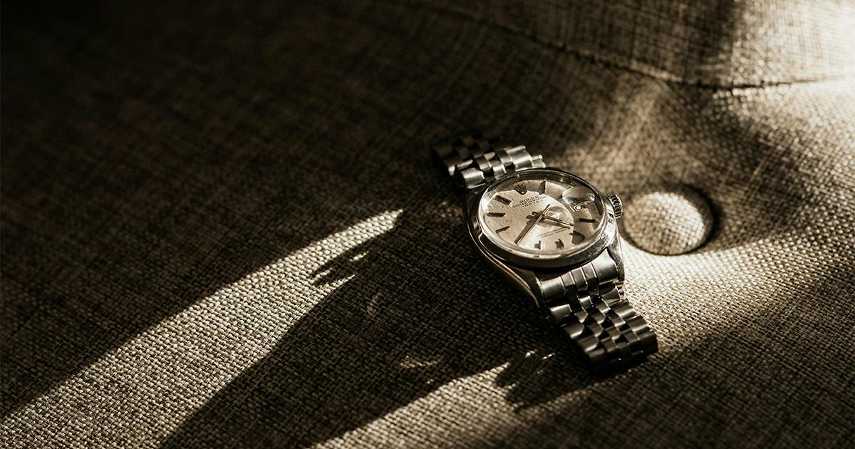 A steel wristwatch with a metal bracelet rests on a textured grey fabric surface, illuminated by a beam of sunlight.