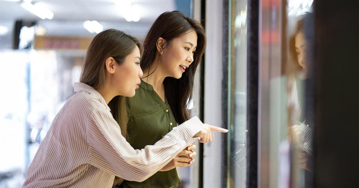 Two young Asian women looking into a luxury retail store window display while pointing at items inside.