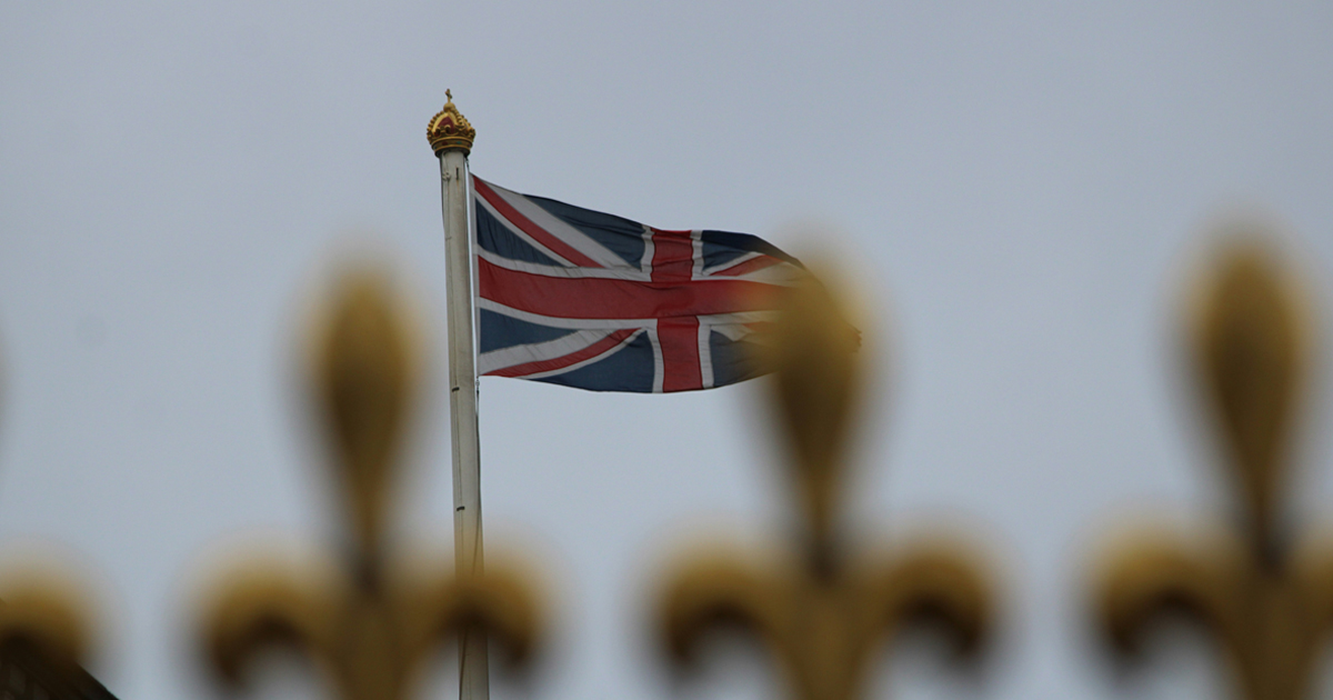 British flag flying on a flagpole, seen behind decorative golden fleur-de-lys railings.