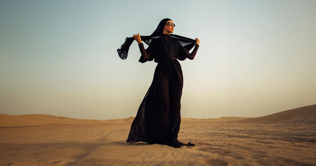 Woman in a flowing black abaya and headscarf standing on desert dunes.
