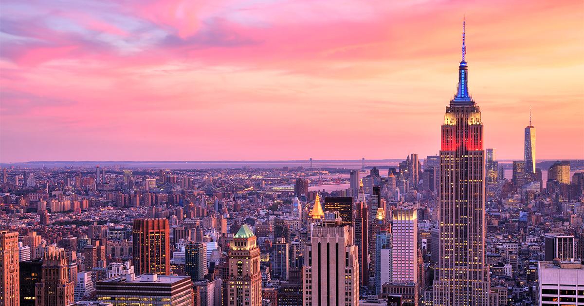 Panoramic view of the New York City skyline and Empire State Building illuminated under a pink and orange sunset sky.