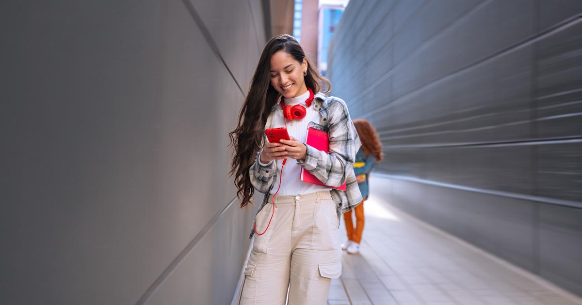 Teenager wearing red headphones smiles while scrolling on her smartphone in a walkway.
